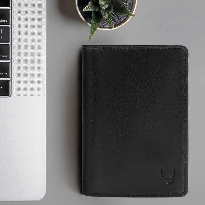 Black leather bi-fold wallet for men, placed beside a laptop and a plant for a modern, minimal look.